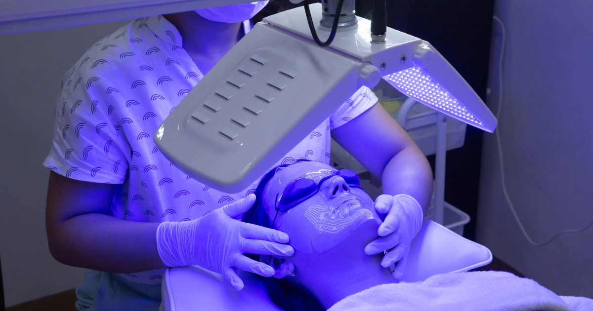A person receiving LED blue light facial therapy with protective goggles as a professional supports their head during LED Light Therapy in Freeland MI.